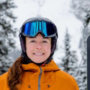 woman in snow gear holding skis smiling at camera with snowy alpine background