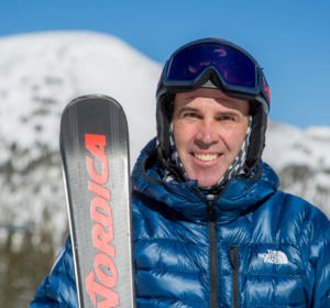 Man in blue coat holding skis with snowy mountain in background