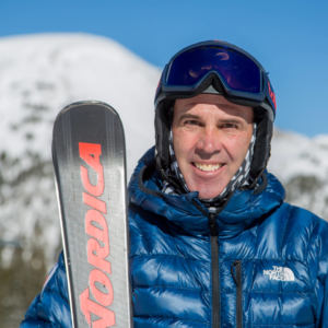 Man in blue coat holding skis with snowy mountain in background