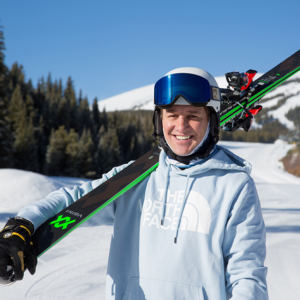 Man in light blue hoodie holding skis with snowy mountain background