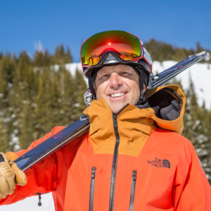 man in orange coat holding skis on shoulder